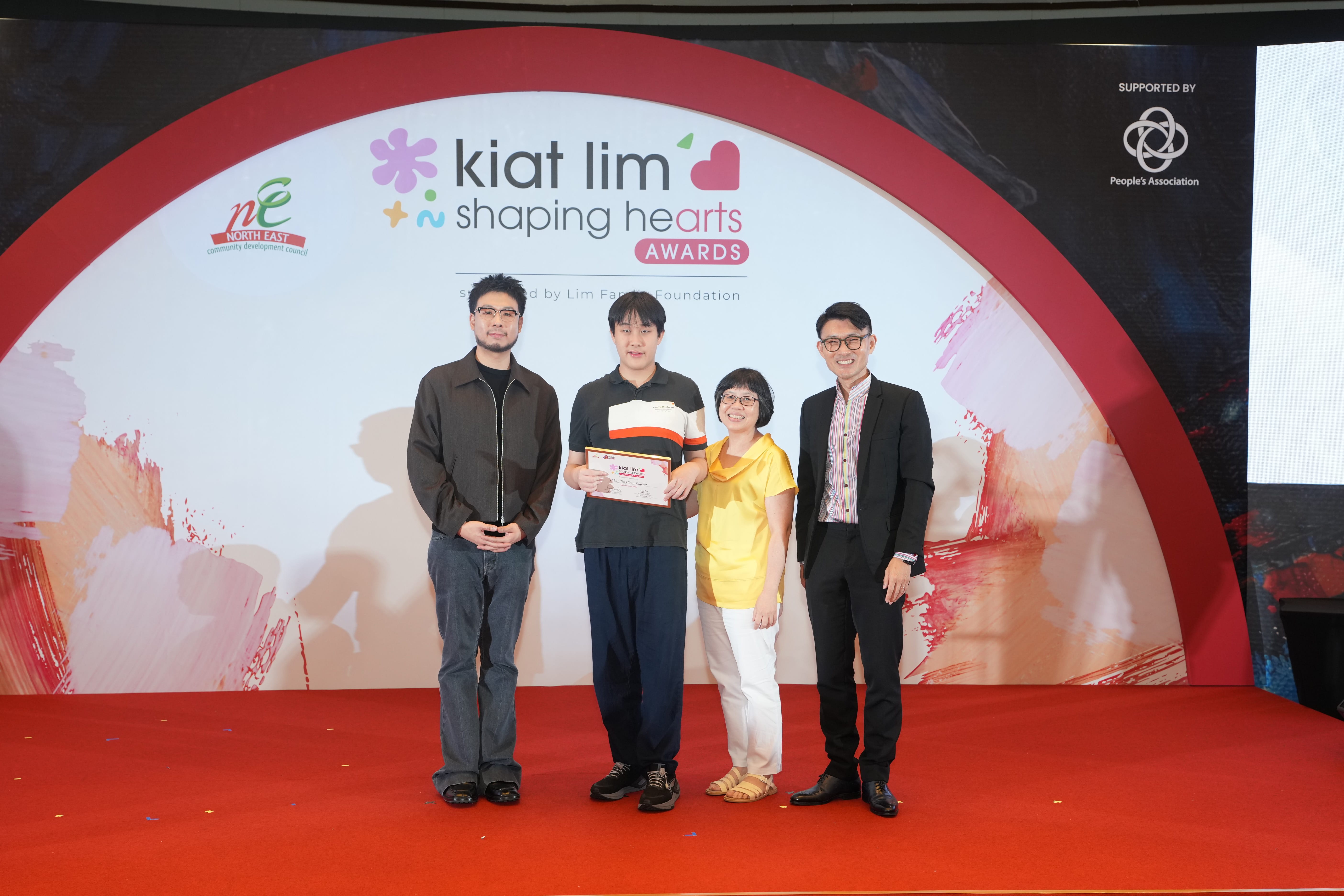 Award recipient standing on stage with the presenter, holding their certificate and posing for a group photo during the award ceremony, with a backdrop reading ‘Kiat Lim-Shaping Hearts Awards'.